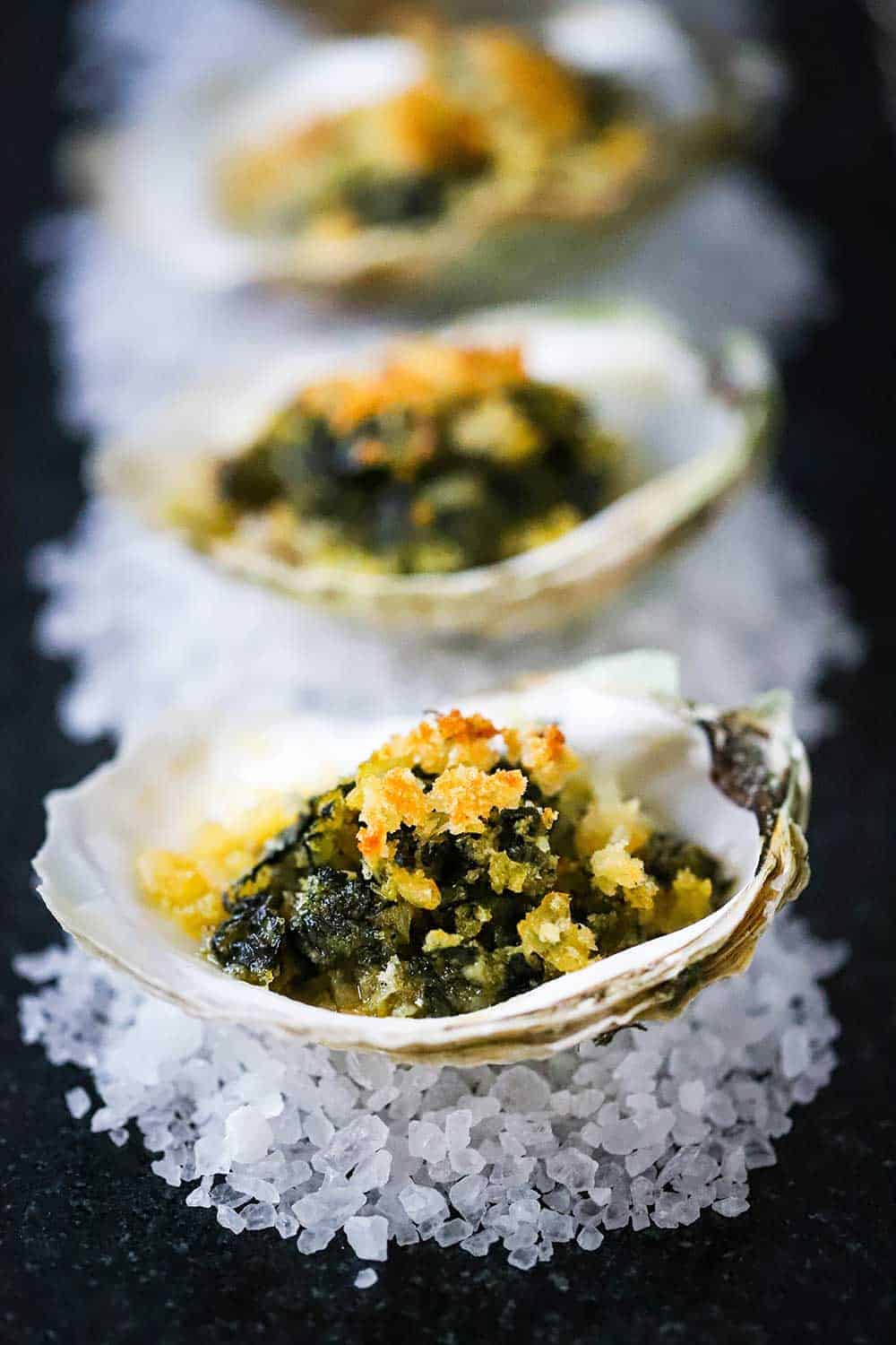 Two oysters Rockefeller each sitting on rock salt on a grey surface with a lemon slice nearby. 