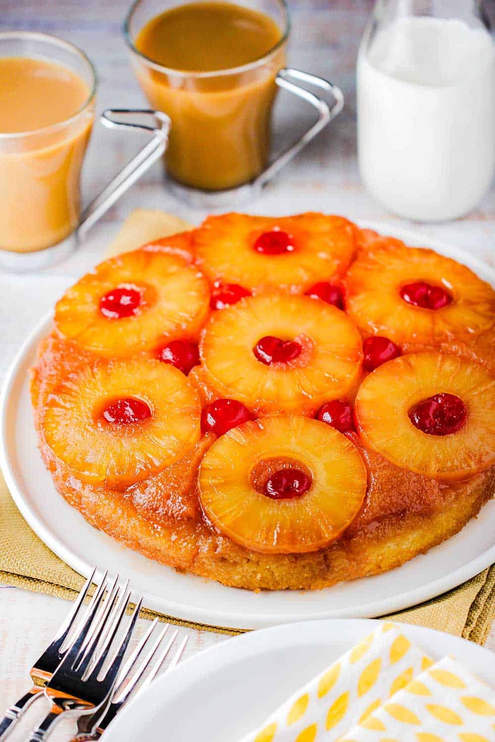 A pineapple upside-down cake on a white platter next to a glass of orange and a cup of coffee.
