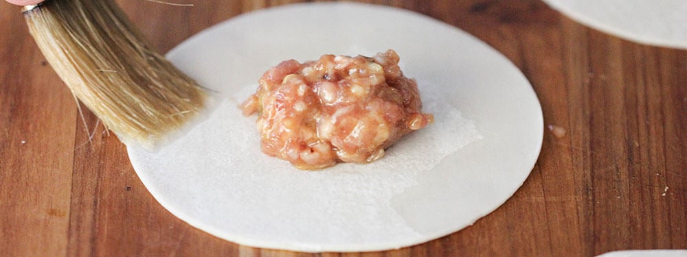 A brush applying water along the edges of a dumpling wrapper for pork dumplings with spicy peanut sauce. 