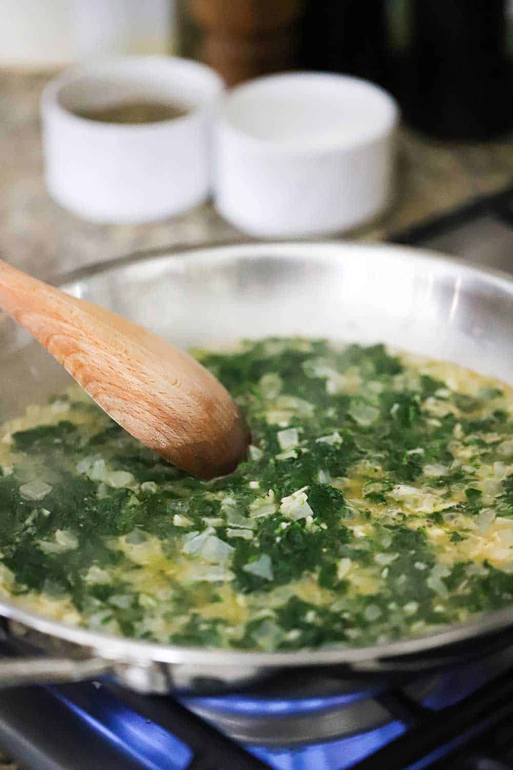 A large silver skillet filled with sautéd spinach, onions, and garlic in a sauce with a wooden spoon in the pan. 