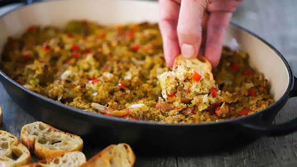 A hand using a slice of toasted baguette to scoop up a helping of baked clam dip. 