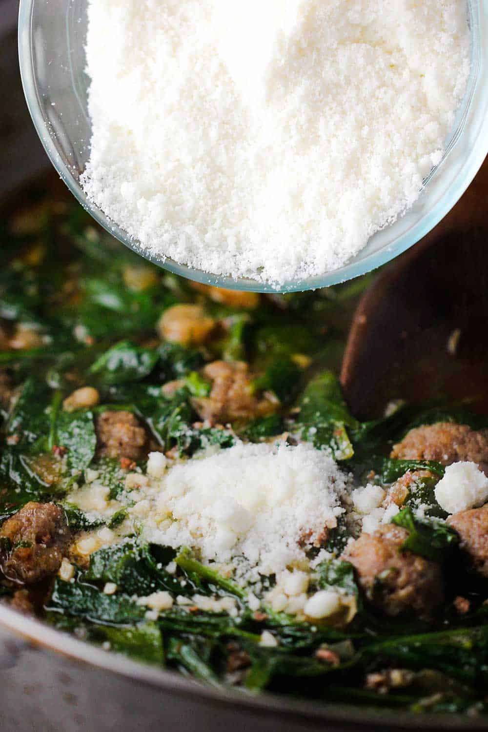 A small bowl of grated parmesan cheese being added to a skillet of pasta sauce with beans and spinach. 
