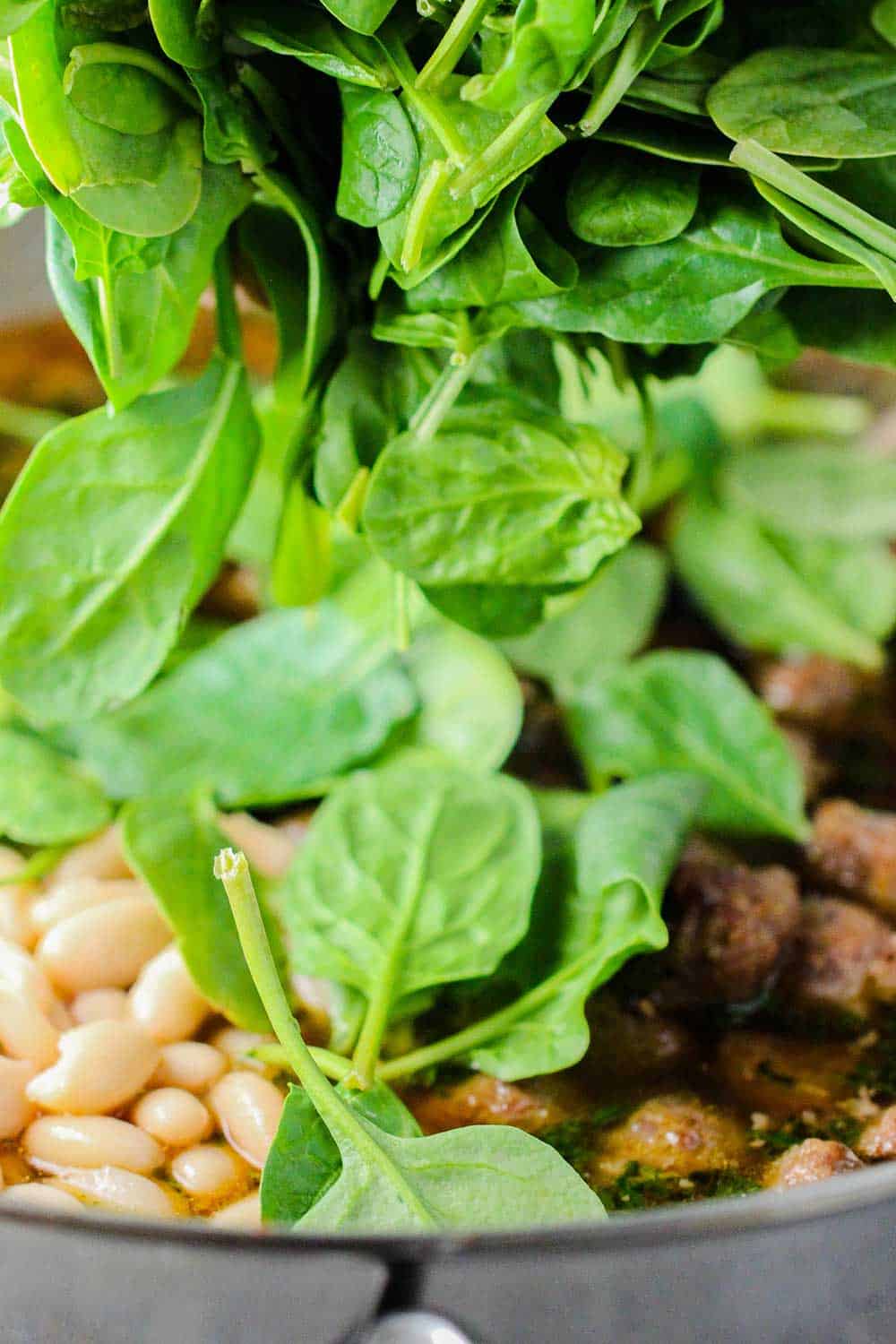 A mound of fresh baby spinach being dropped into a skillet of country Italian pasta sauce. 