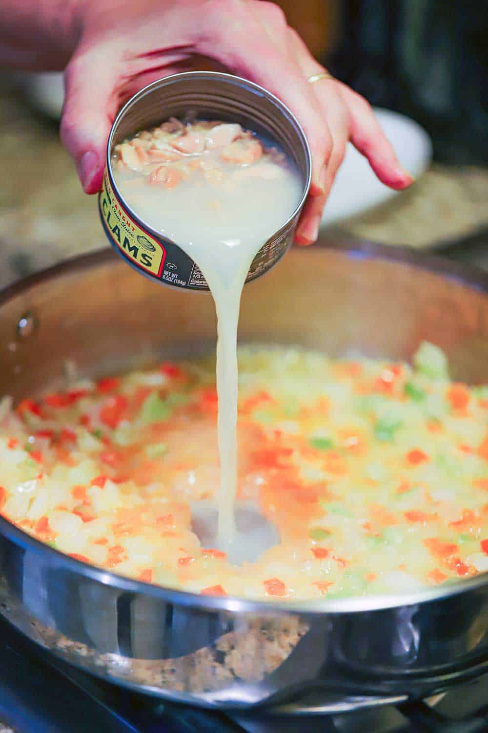 A hand pouring canned clams with their juice into a silver skillet filled with sautéd vegetables. 