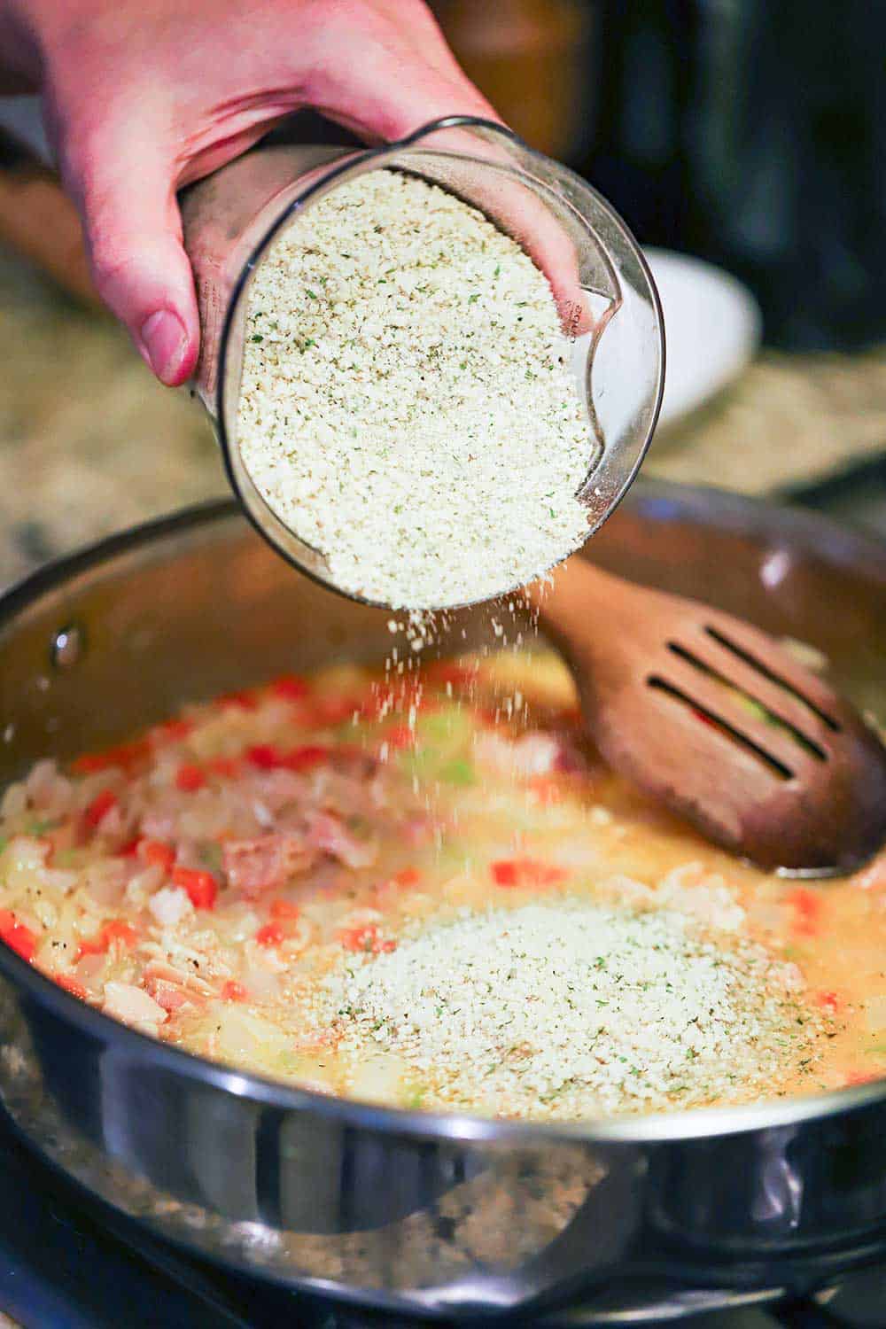 A hand dumping Italian-style Panko breadcrumbs into a skillet filled with clams, clam juice, and sautéd vegetables. 