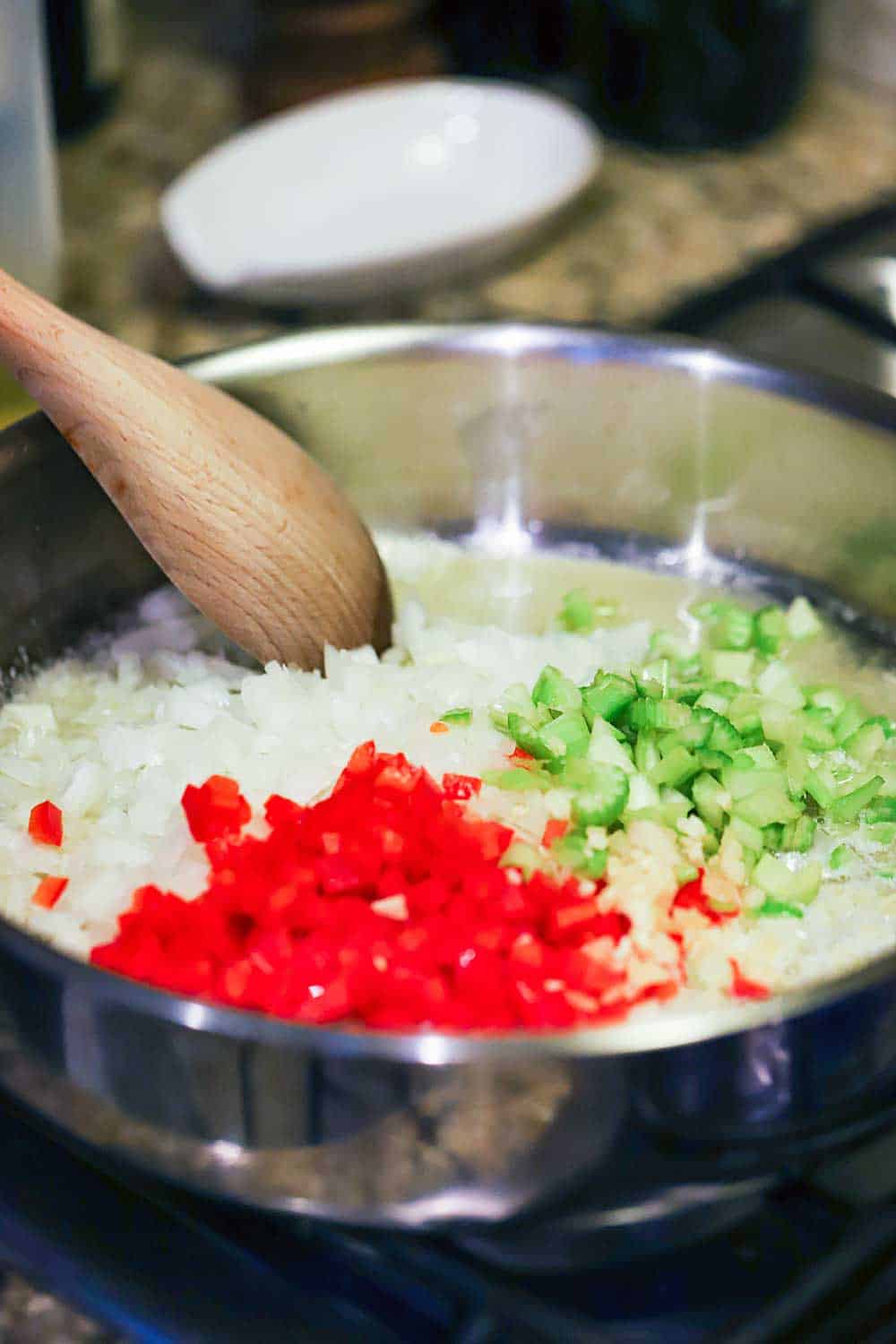 A large stainless steel skillet filled with chopped onions, red bell peppers, and celery, all being stirred by a large wooden spoon. 