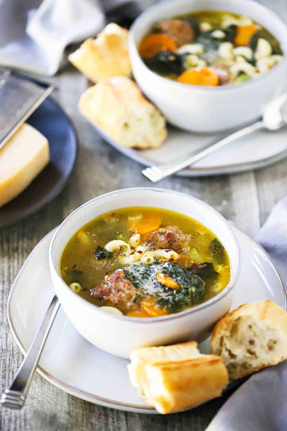 Italian Wedding Soup in a white bowl on a small white plate with a couple of pieces of bread on it.