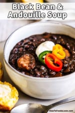 A close-up view of a soup bowl filled with black bean and andouille sausage topped with sliced peppers and a dollop of sour cream on a plate with a piece of cornbread on it.