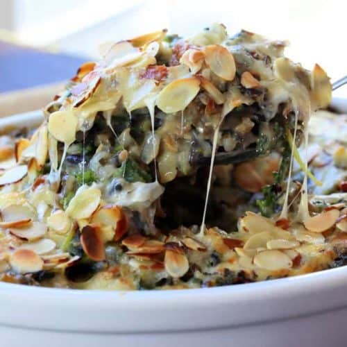 A spoon lifting our a helping of Wild rice, kale, and fontina casserole in a white dish.