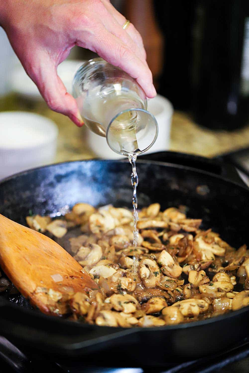 A hand pouring white wine from a small carafe into a cast-iron skillet filled with sautéd sliced mushrooms. 