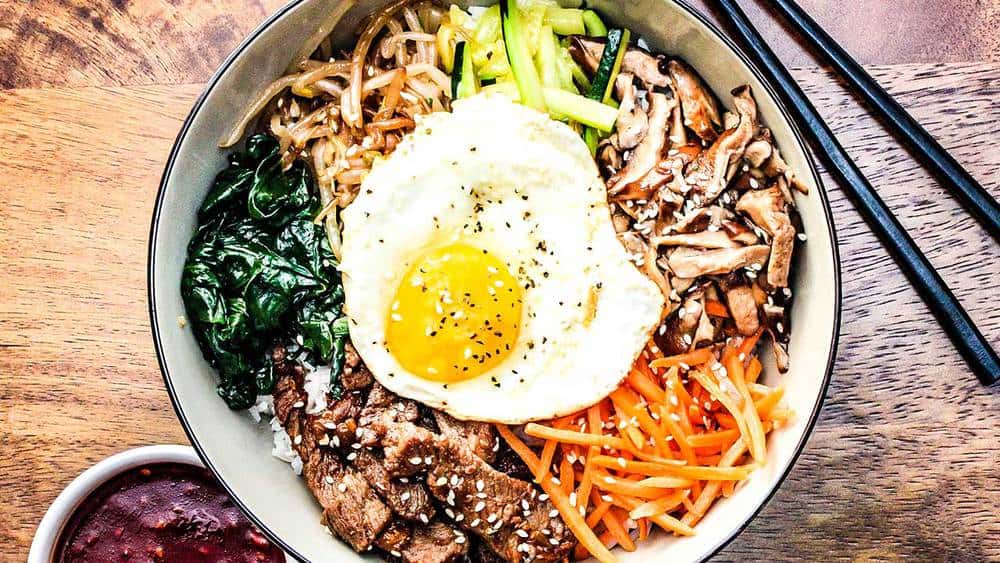 A bowl of authentic bibimbap next to Korean BBQ sauce and chop sticks