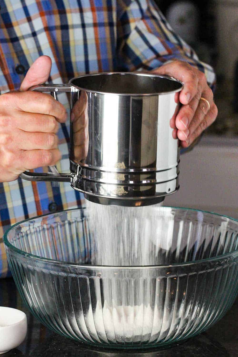 Two hands holding a large sifter while sifting flour into a glass bowl. 