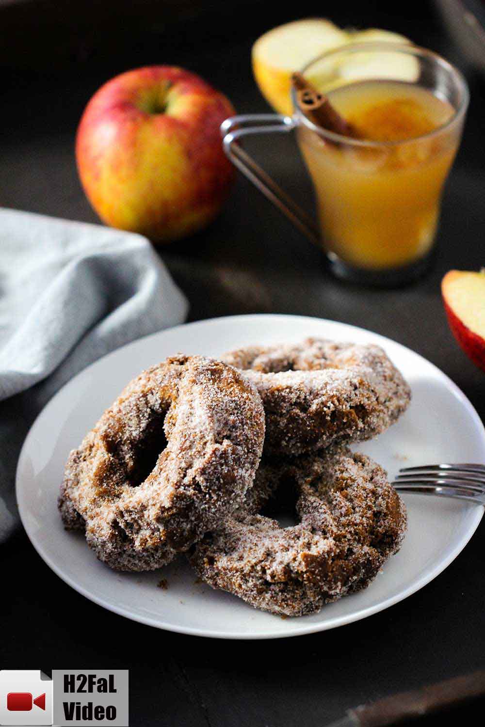 A white plate with three apple cider doughnuts on it with an apple and a glass of cider next to it. 