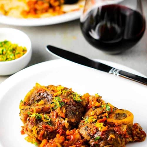 A white plate with the dish Osso Buco on it next to a glass of red wine