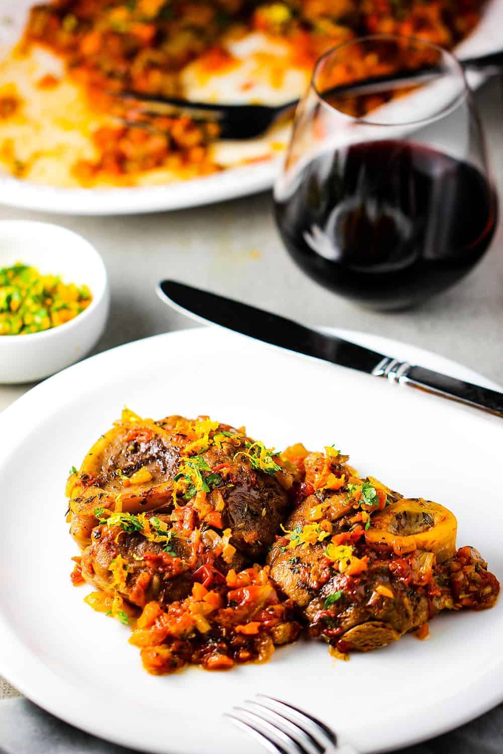 A white plate with the dish Osso Buco on it next to a glass of red wine