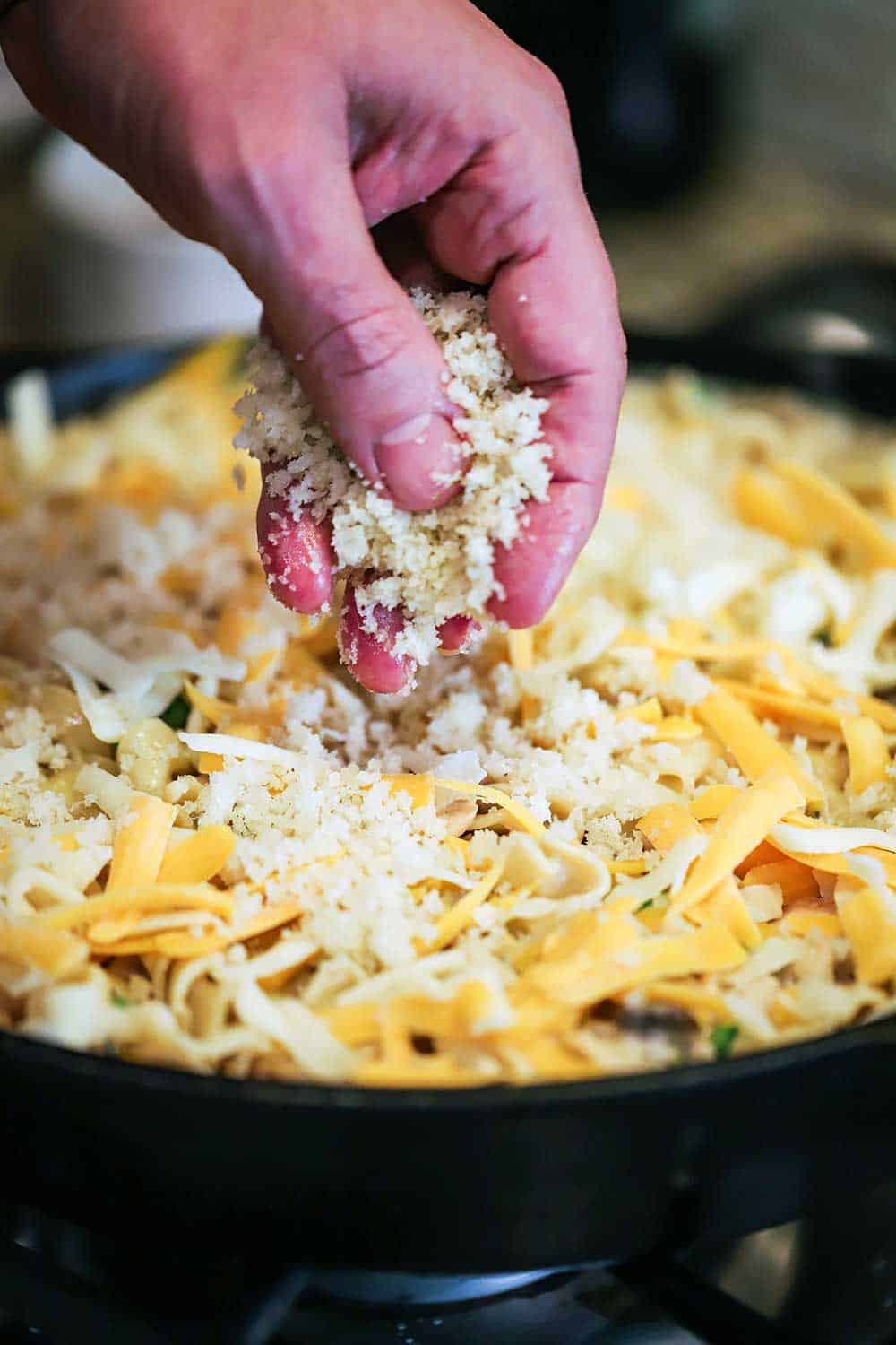 A hand sprinkling homemade bread crumbs over the top of a tuna casserole in large cast-iron skillet. 