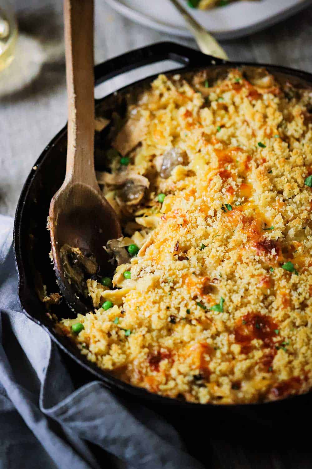 A large black cast-iron skillet filled with tuna casserole with a serving missing from the pan and a wooden spoon inserted in the side of the pan. 