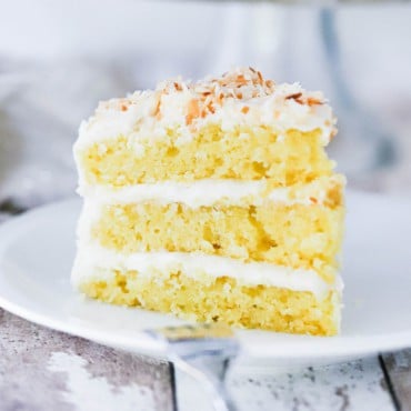 A 3-layered slice of coconut cream cake standing upright on a plate in front of the cake on a cake stand.