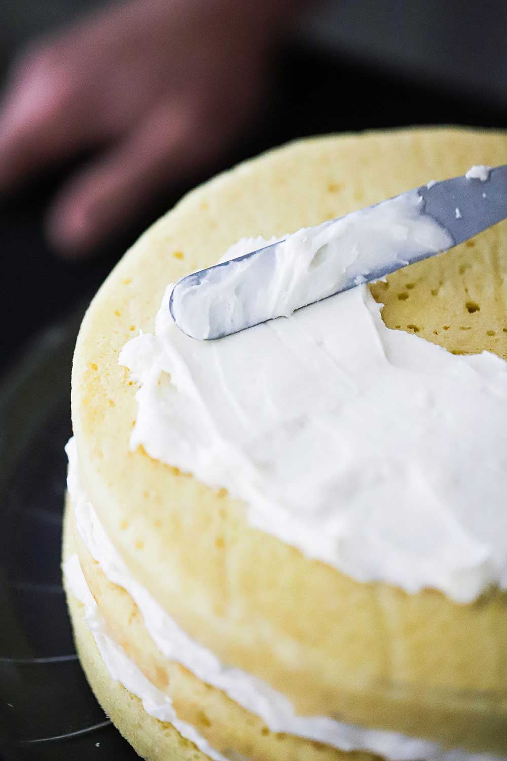 An off-set spatula smearing cream cheese frosting on a 3-layered yellow cake. 