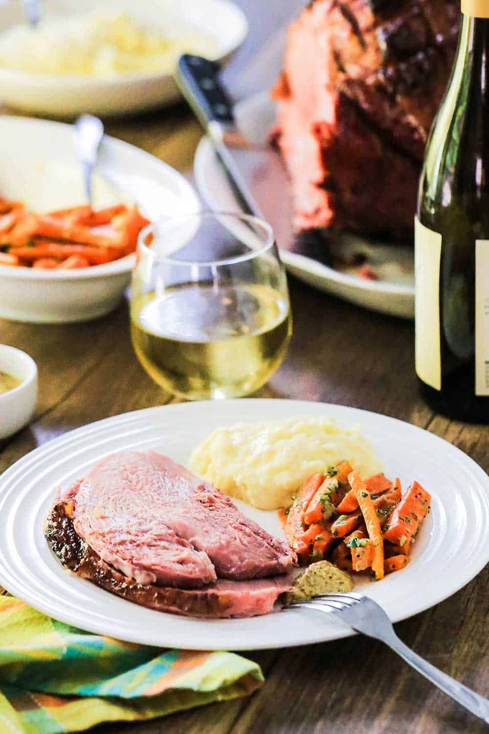 A white dinner plate with sliced ham, glazed carrots and mashed potatoes on it, next to a glass of white wine. 