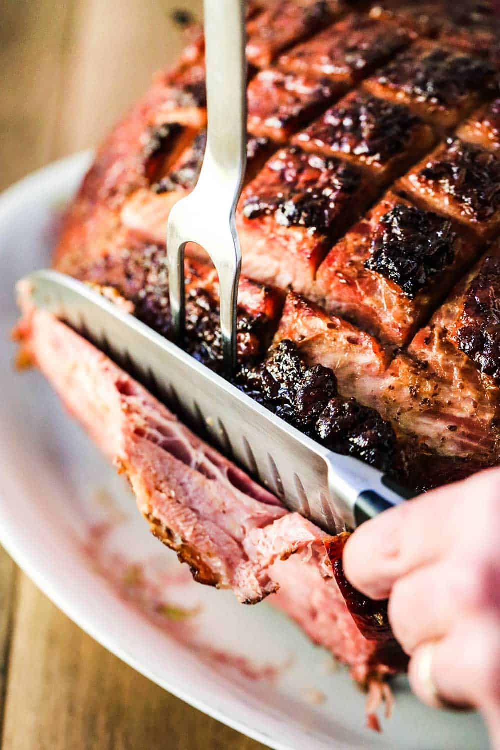 Two hands using a large fork and knife to carve a honey bourbon glazed ham on a platter. 