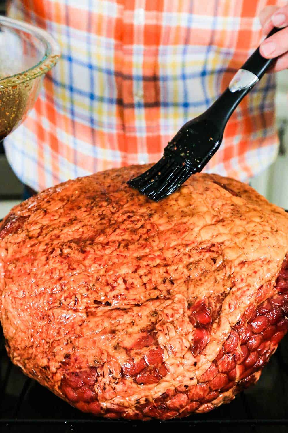 A hand using a large brush to apply glaze to an uncooked Berkshire ham. 