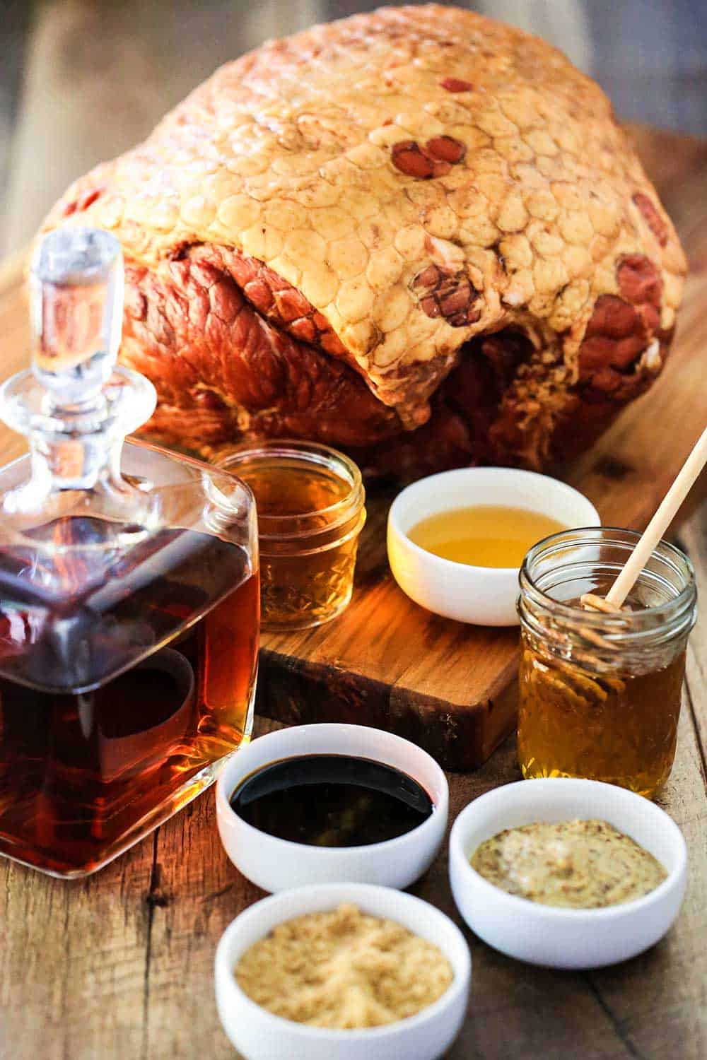 A cutting board with a large Berkshire ham in the background and a sniffer of bourbon, and small bowls of apple cider vinegar, mustard, and brown sugar next to it. 