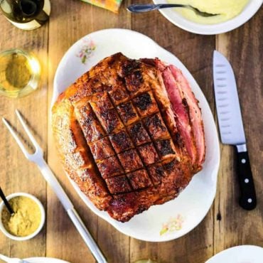 An overview shot of a honey bourbon glazed ham next to a bowl of mashed potatoes, glazed carrots, and a glass of white wine.