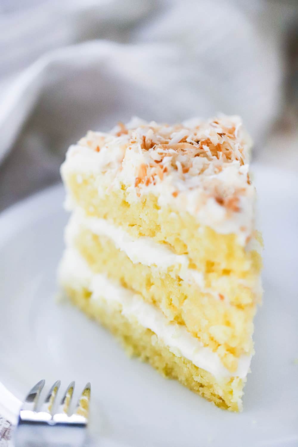 A 3-layered slice of coconut cream cake on a white dessert plate. 