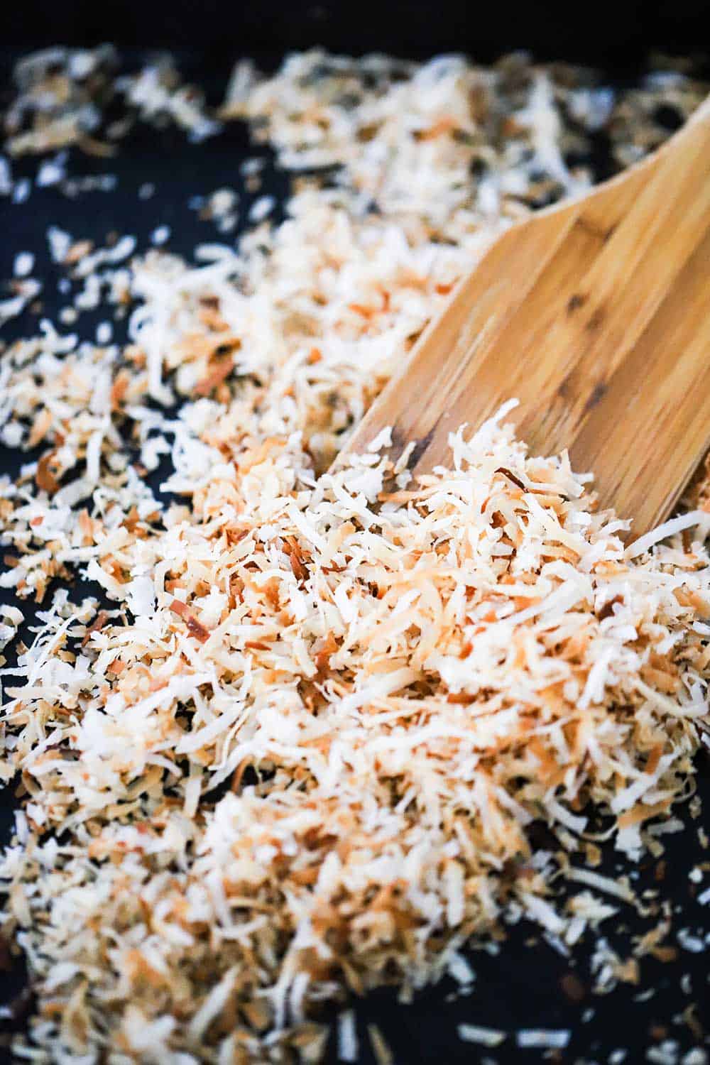 A black baking sheet filled with toasted coconut flakes that are being scooped up from a wooden spatula. 