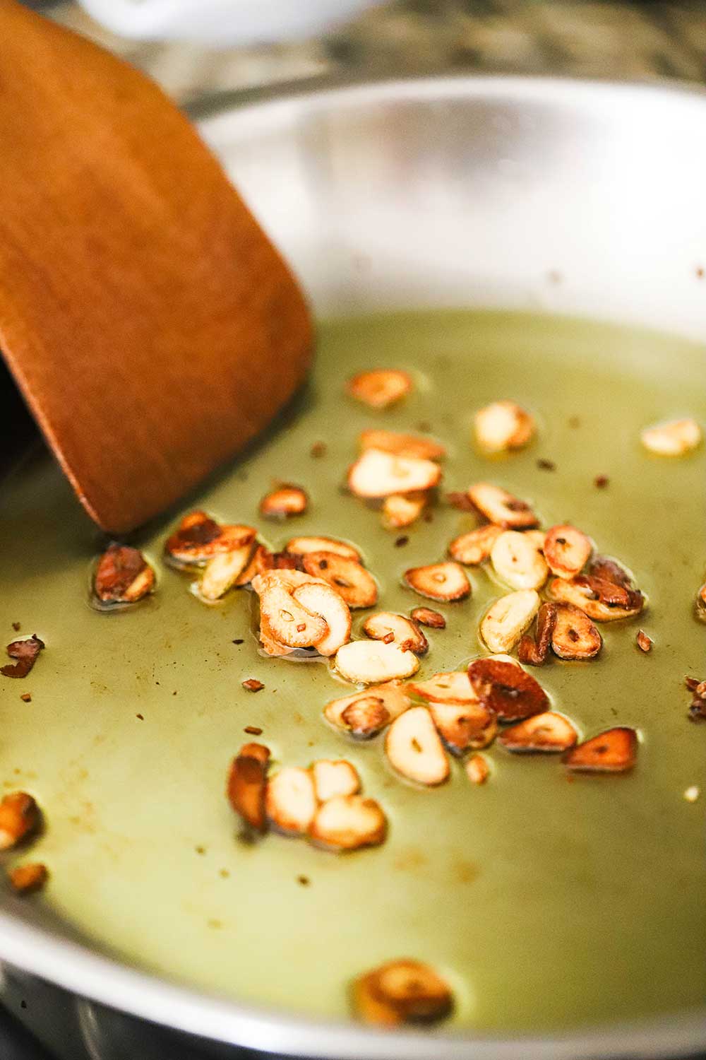 A large silver skillet filled with simmering olive oil and garlic slivers that have turned a golden brown, with a wooden spoon stirring them in the pan. 