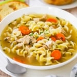 A white soup bowl filled with chicken noodle soup sitting on a dinner plate with a soup spoon tucked in the side and toasted bread slices nearby.