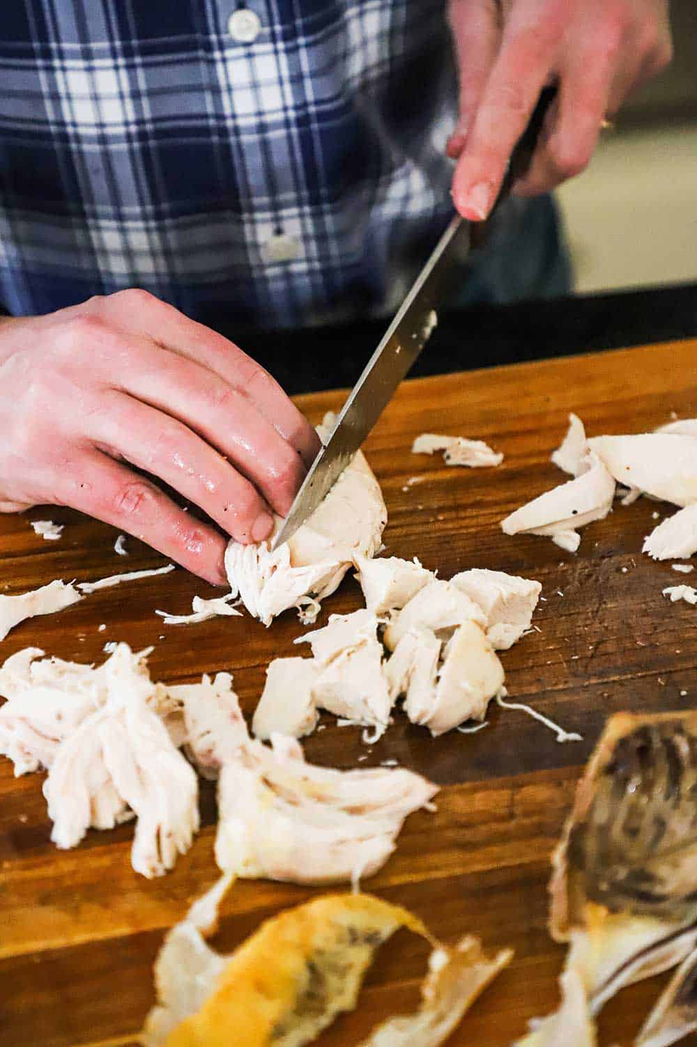 A person using a chef's knife to cut up cooked chicken breasts on a wooden cutting board. 