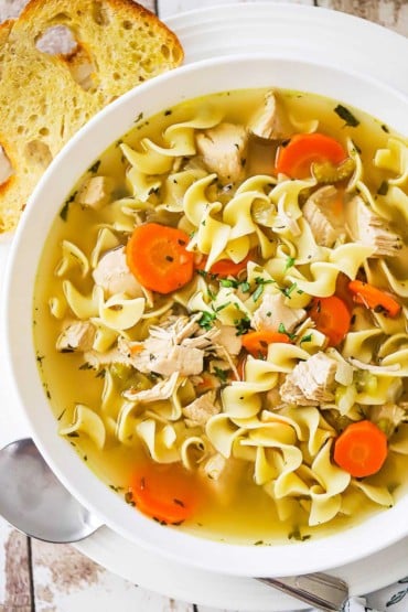 A white soup bowl filled with homemade chicken noodle soup next to a sliced of toasted bread.