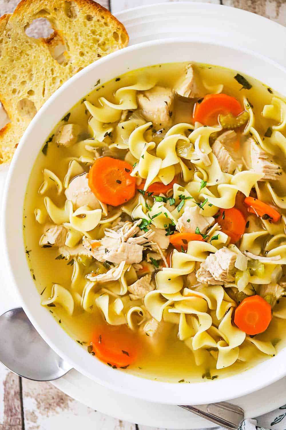 A white soup bowl filled with homemade chicken noodle soup next to a sliced of toasted bread. 