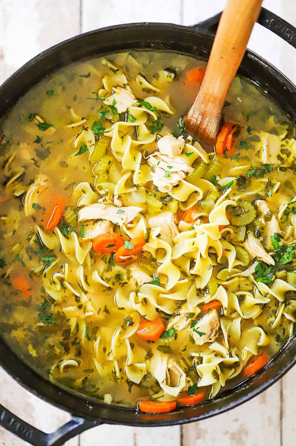 A black pot with handles filled with homemade chicken noodle soup topped with chopped fresh parsley. 