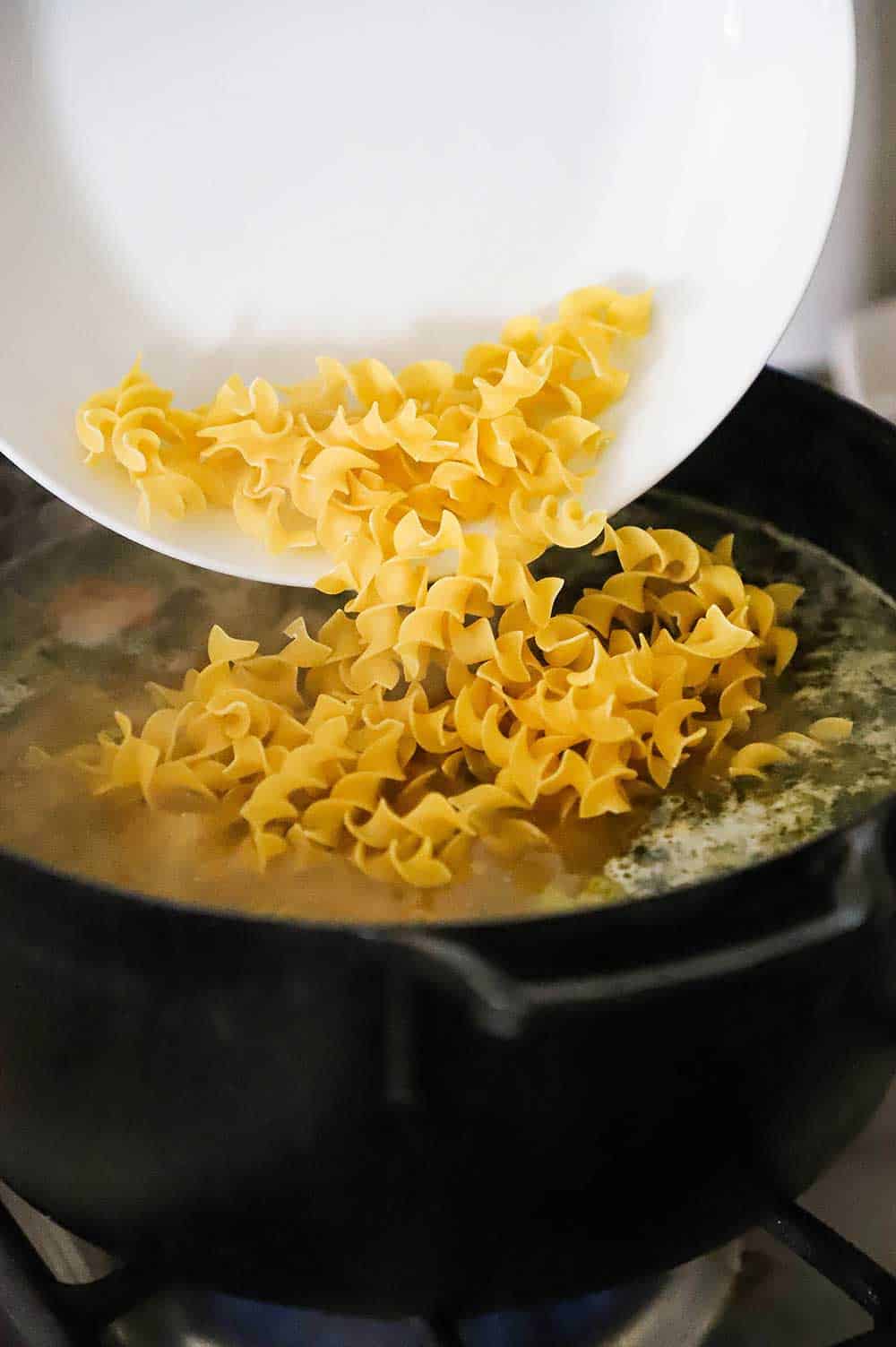 Uncooked pasta egg noodles being transferred from a white bowl into a black pot filled with simmering broth and cooked vegetables. 