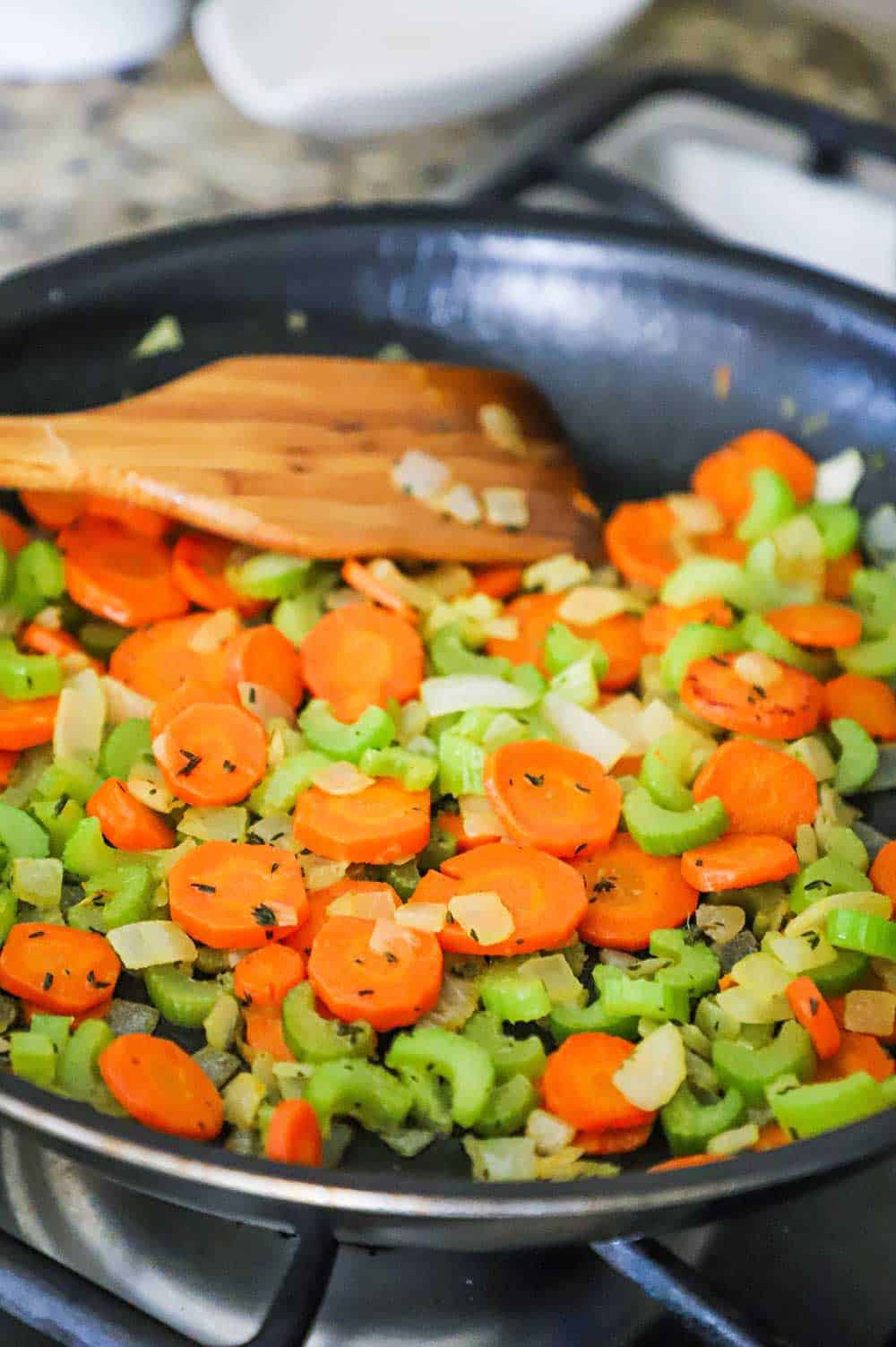 A large non-stick black skillet filled with sliced carrots, celery, and chopped onion with a wooden spoon inserted in the side. 