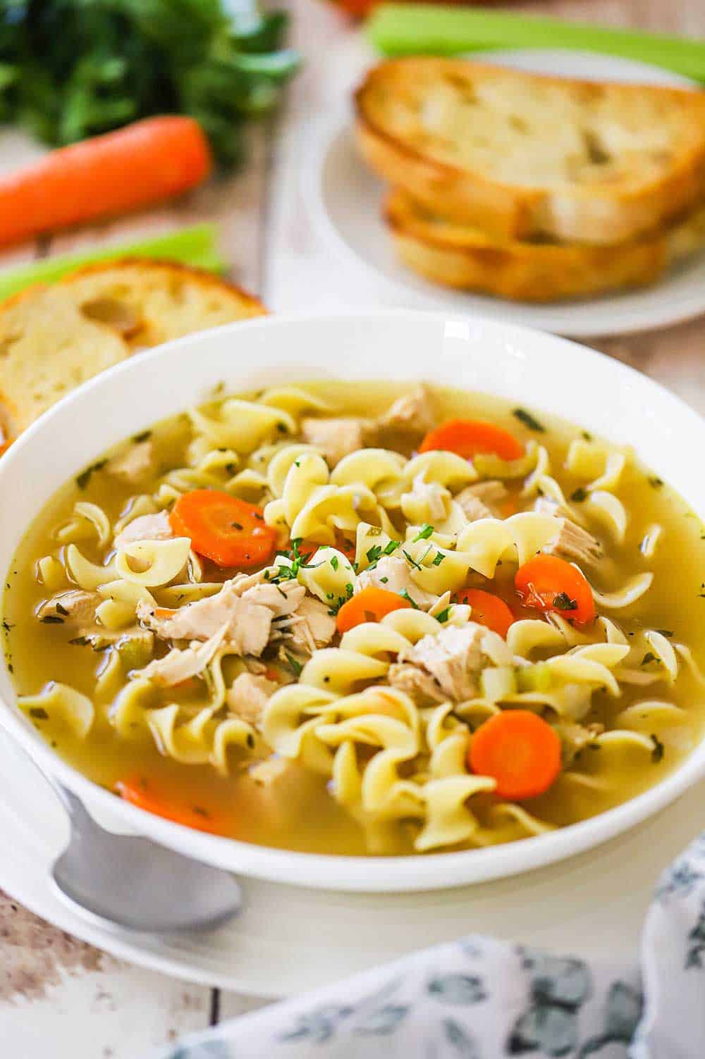 A white soup bowl filled with chicken noodle soup sitting on a dinner plate with a soup spoon tucked in the side and toasted bread slices nearby. 