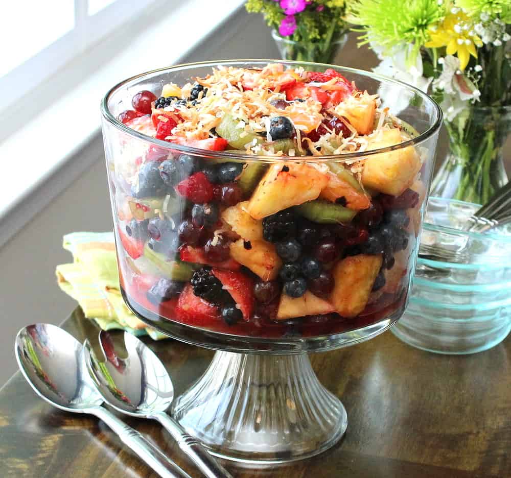 A glass trifle filled with a summer fruit salad topped with toasted coconut sitting on a table near a window.
