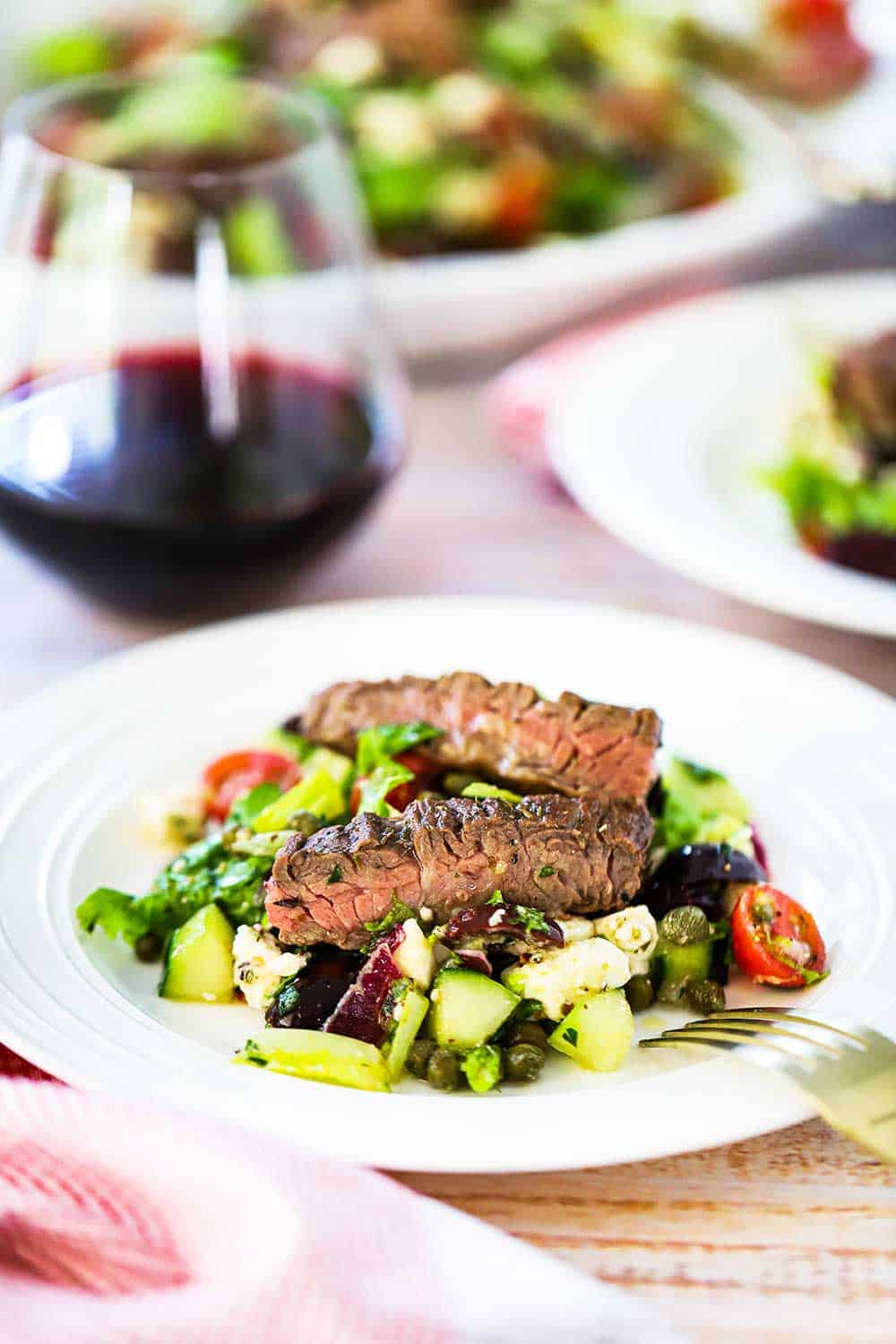 An individual plate filled with Greek salad with grilled meat sitting next to a glass of red wine. 