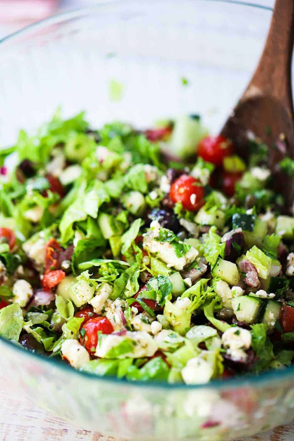 A glass bowl filled with a chopped Greek salad with a wooden spoon stuck in the middle. 