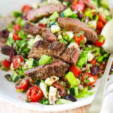 A large oval platter filled with a Greek salad and grilled steak with two large gold spoons on the side.