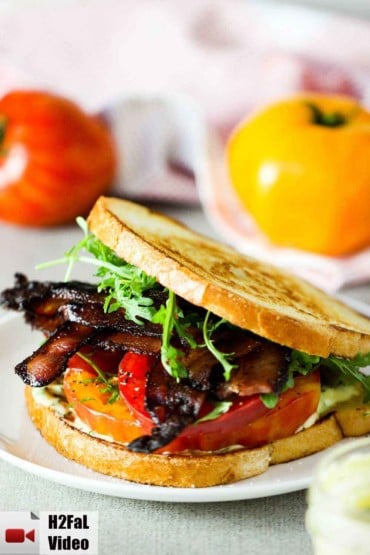 A classic BLT sandwich with garlic aioli on a white plate with two tomatoes in the background.