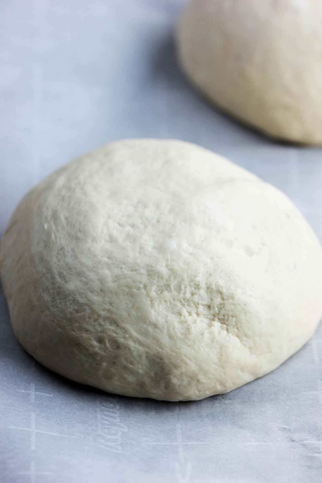 A ball of semolina pizza dough sitting on parchments paper next to another dough ball.