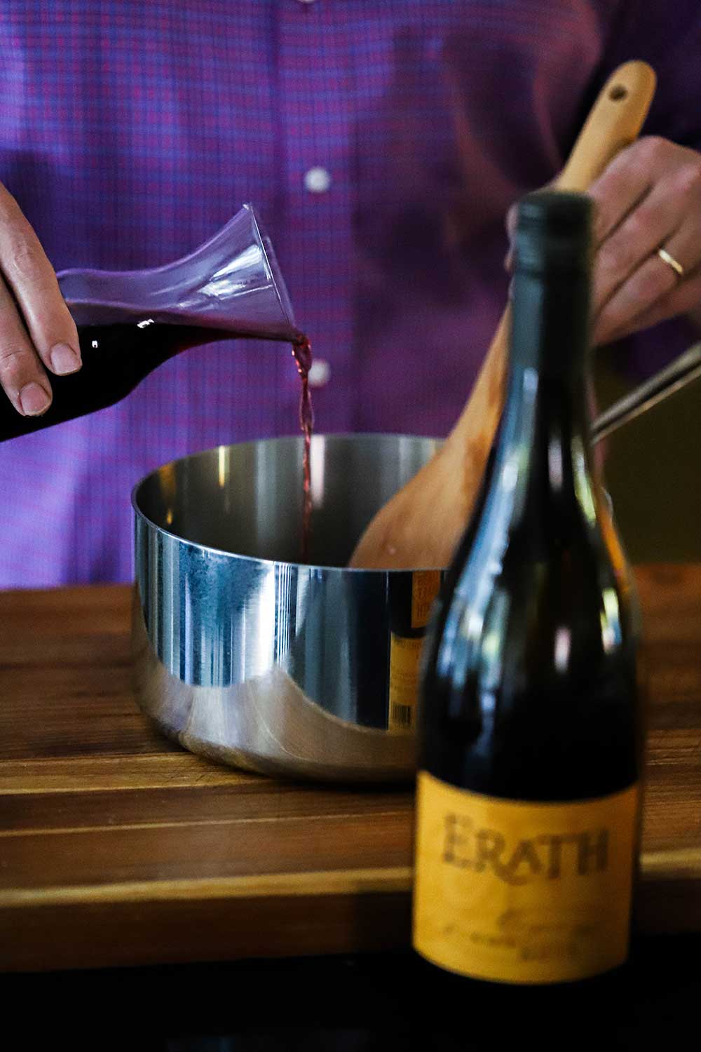 A person pouring red wine from a carafe into a silver sauce pan all in front of a bottle of Erath Pinot Noir.