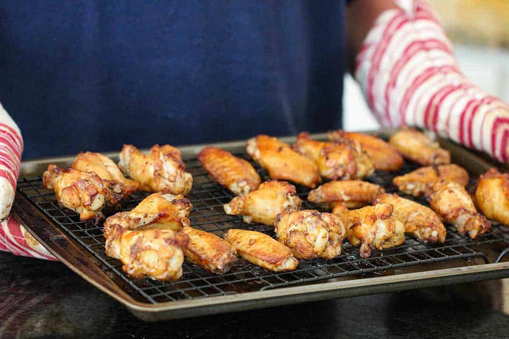 Two hands with oven mitts holding a baking sheet of baked chicken wings. 