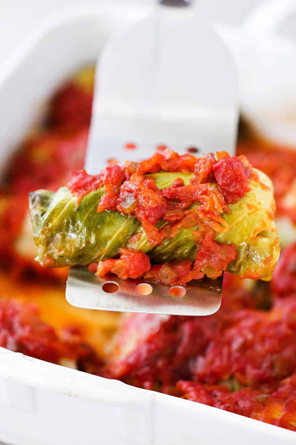 A metal spatula holding up a stuffed cabbage roll over a dish of the same. 