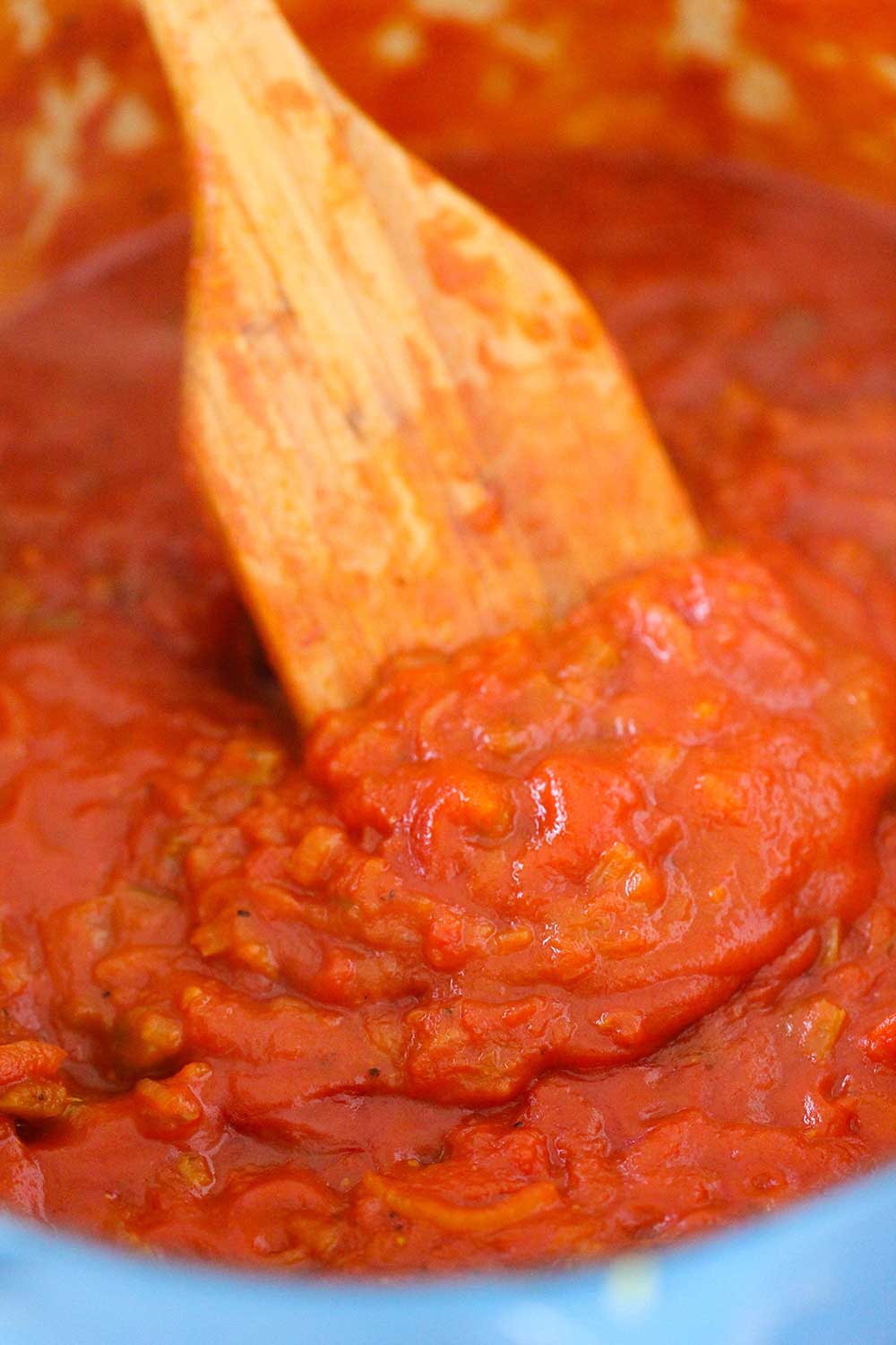 A wooden spoon stirring tomato sauce in a blue Dutch oven. 