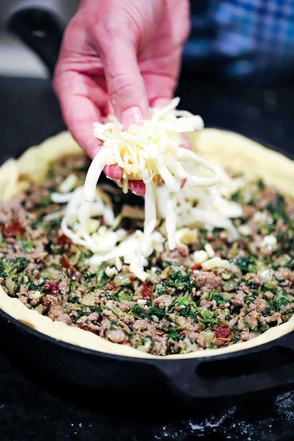 A hand sprinkling shredded mozzarella cheese over the top of deep dish pizza with a sausage and spinach filling all in a cast-iron skillet. 